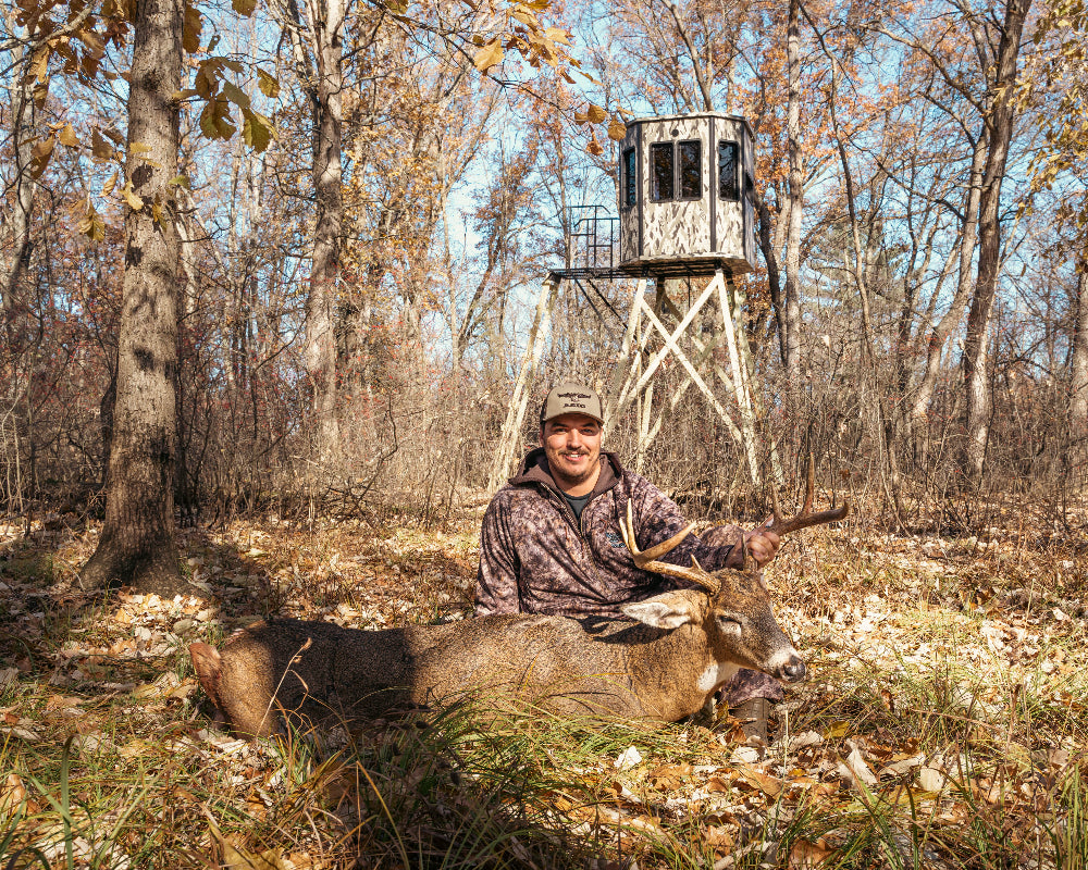 Man with a deer and hunting rifle in front of a modular blind on wood and metal tower in a forest.